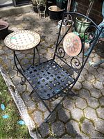 Outdoor bistro chair and table on a brick patio. The chair is black metal with lattice seat and a decorative round tile inlay on the backrest. The table is round with a mosaic tile top in beige and white.