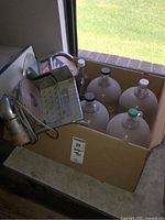 Photo showing six large glass bottles in a cardboard box with a vintage manual meat slicer leaning on the box near a window.