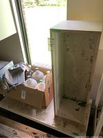 Photo showing the wooden cabinet with visible stains and wear, located next to a window with items around it (contents not included).