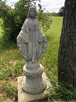 Full view of the cement Virgin Mary statue standing outdoors on pedestal base surrounded by grass and tree.