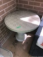 Full view of round cement side table placed in corner against brick wall, showing pedestal base and textured tabletop.