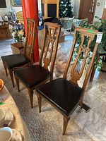 Front and side view of three wood dining chairs with black faux leather seats and detailed carved backs. Two captain chairs have broken arms. Set shows finish wear and nicks.