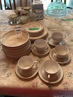 Full view of Alfred Meakin Gold Crown plates, bowls, Royal Knight cups and saucers arranged on a lace tablecloth beside a glass cake dome.