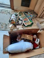 Box containing assorted Christmas ornaments including figurines, batting, and decor items.