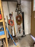 Photo showing several shovels, orange and black extension cords, a metal pipe against a basement wall near electrical outlets and a light fixture.