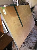 Large wooden drafting table leaning against a wall, stained and marked, green metal adjustable support brace visible.