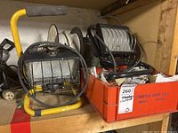 Two black portable work lamps with yellow foam padded handle grips and protective metal grilles on the lens, surrounded by wire reels and electrical components.