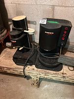 Two black Faema Family coffee makers placed side by side on a wooden surface against a concrete wall, showing overall condition and size.
