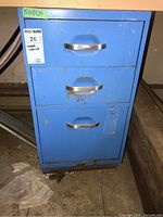 Front view of blue metal filing cabinet showing three drawers with metal handles, scratches, rust and wear visible on surface.