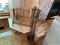 Brushed brass fireplace log holder and set of fireplace tools standing beside a marble hearth, showing overall condition and size.