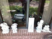 Four white garden statues and the mirror panel arranged on a brick ledge in front of a window. Statues depict cherubs and children figures.