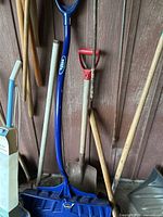 Various shovels including blue snow shovel, wooden handle shovel with red grip, and others leaning against a wall
