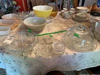 Wide view of assorted glass and ceramic servingware laid out on lace tablecloth including bowls, glass cake dome with base, ashtrays, trinket box, and plates.
