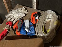 Photo showing white work gloves, orange and blue ratchet tie down straps, white towing rope, and red fabric protective sleeve placed on a wooden surface next to a cardboard box.