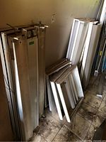 Group of multiple light fixtures stacked against a wall on a dirty floor. Includes long rectangular and square panels, with visible wiring and mounting hardware.