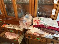 Wide view of three drawers from buffet and top surface showing folded linens such as tablecloths, placemats, napkins, and doilies with some fabric rolls and stacks.