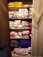 Six shelves of assorted folded linens, towels, blankets including a rolled pink and white fabric, blue fabrics and others.