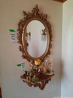 Photo showing the oval ornate wall mirror with detailed carved gold wood frame, also showing the matching wall shelf under it with decorative items on top.
