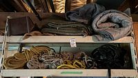 Top shelf with folded moving blankets in gray, green, and tan along with coils of various ropes in natural and synthetic fibers.