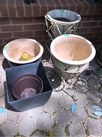 Four plant pots arranged together outdoors on stone tile floor; includes two large cream ceramic pots (one with metal stand), smaller cream ceramic pot, and two plastic pots (round brown, square black).