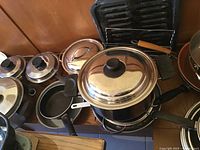 Photo showing multiple pots and pans with lids, several with black handles, a roasting sheet, and a grill rack in the background.