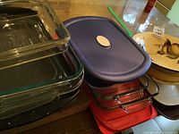 Stack of Pyrex glass dishes with purple and red resin lids alongside ceramic fruit bowl and other kitchen items on table