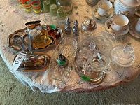 Overview of metal and glass servingware items on lace-covered table including trays, salt and pepper shakers, glass cream and sugar sets, and spoons.