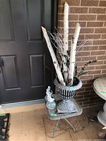 Outdoor side table with decorative plastic plant pot containing faux birch branches and two angel solar lights on top, placed next to a front door and brick wall.