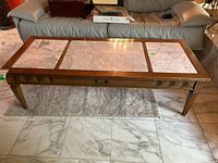 Front view of long rectangular wood coffee table with three rectangular marble insets on top, light marble with gray veining, placed on a rug in front of a leather sofa.