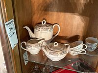 View of the tea set including teapot, sugar bowl, creamer, and cups with saucers on wooden shelf, showing overall set details and condition.