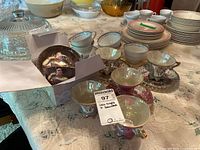 Multiple teacups and saucers arranged on lace tablecloth, including iridescent and floral designs