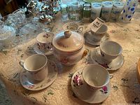 Collection of teacups, saucers, and covered bowl arranged on table with lace tablecloth. Some teacups have floral patterns and gold trims.