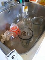Photo showing 8 different glass vases placed inside a kitchen sink, including a ribbed vase, a vase with handle, a jar with orange filler, and a vase with yellow stopper.