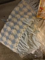 Close-up of light blue and cream crocheted bunting bag with tassel fringe edges laid out on floor.