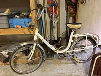 Side view of the white vintage folding bike showing the frame, tires, seat, pedals, and rear rack in a basement workroom.