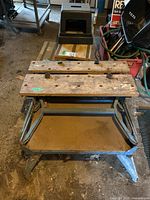 View of folding metal work bench with wooden slats showing holes and clamps, with black plastic step stool on top in garage setting.