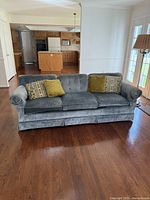 Full frontal view of medium blue plush fabric couch with three cushions, two decorative pillows, traditional rolled arms and skirted base, situated on hardwood floor with kitchen background.
