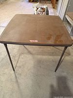 Photo of a large laminate card table with folding metal legs and a vinyl top showing surface wear and stains on concrete floor