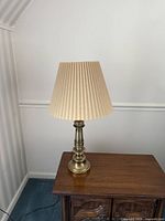 Single patinated brass table lamp with beige pleated shade on wooden side table, against a white wall