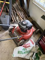 Red Scott lawn mower with a black 6.0 HP engine sitting on a floor near wood pieces and a window. A grass collection bag is partially visible in front of the mower.
