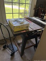 Full view of the Sears Craftsman table saw on black metal stand with box and plastic bag on top, positioned near window.