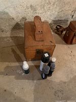 Wooden shoe shine box with handle and metal latch on concrete floor. Multiple shoe care product bottles placed in front of the box.