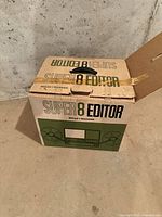 Closed original box of Super 8 Editor, beige cardboard with green and black text and graphics, labeled 'Super 8 Editor Atlas + Warner'. Box shows some wear and taped repairs.