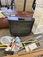 Photo taken showing a black Sony CRT television with remote control on top, surrounded by various cleaning tools and miscellaneous items in a cluttered room environment.