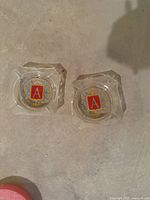 Top-down view of both vintage glass ashtrays showing the red 'A' emblem on shield and wreath design in the center.