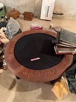 Miniature trampoline sitting on a basement floor surrounded by various items, showing top view with brown padded rim and black jumping surface with exercise use wear.