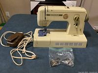 Front angle of Bernina Zigzag sewing machine with power pedal and set of accessories in plastic bag on table.