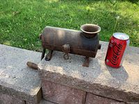 Side view of the cast iron grinder next to a soda can for scale, showing cylindrical body, hopper, hand crank, and three legs with one leg broken.