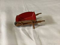 Red metal and wood miniature wheelbarrow lying on a white fabric background, viewed from side angle showing detail of the wooden handles and black wheel.