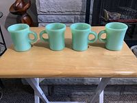 Front view of four light green Jadeite Fireking coffee mugs with C-shaped handles displayed on a wooden table.